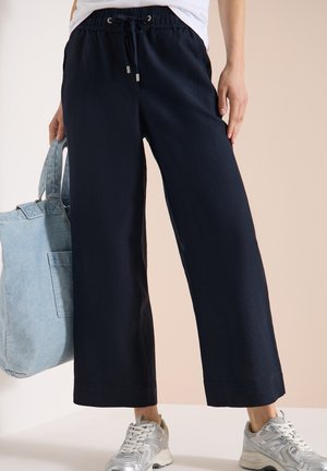 Personne portant un pantalon noir ample et des baskets blanches, tenant un grand sac cabas bleu clair à côté d'elle.