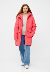 Lange roze parka met een trekkoord in de taille, ritssluiting, twee voorzakken en een capuchon, gedragen over een lichtgrijs sweater en blauwe spijkerbroek.