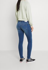 Žena v světle zelené mikině, modrých skinny džínách a černo-bílých teniskách, stojící s výhledem od bílé pozadí.