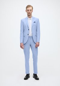 Hellblauer, taillierter Anzug mit einreihiger Jacke, schmal geschnittenen Hosen, weißem Hemd und schwarzen Dressschuhen. Strukturierter Stoff, minimalistische Details.