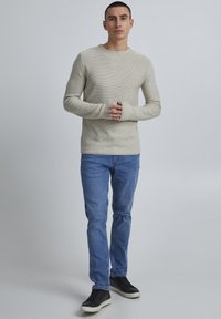 Beige geripptes Langarmshirt, hellblaue Jeans und schwarze Sneakers. Das Shirt hat Daumenlöcher an den Bündchen. Einfaches, lässiges Outfit.