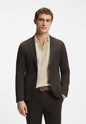 Ung mand står iført mørkebrun blazer, lysebeige strikket polo, brun bælte og matchende mørke bukser mod hvid baggrund.