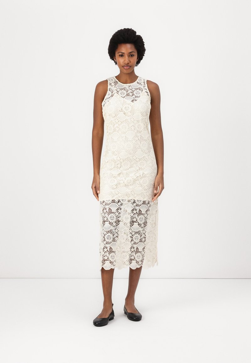 Kanten jurk in crème met een mouwloos ontwerp, gegolfde zoom en bloemmotieven doorheen. Gecombineerd met zwarte platte schoenen.