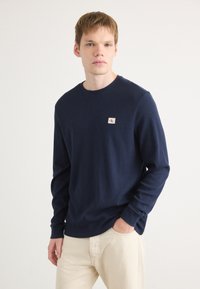 Navyblauwe lange mouwen shirt van een gestructureerde stof, ronde halslijn en een klein logopatch op de linker borst, gecombineerd met crèmekleurige broek.