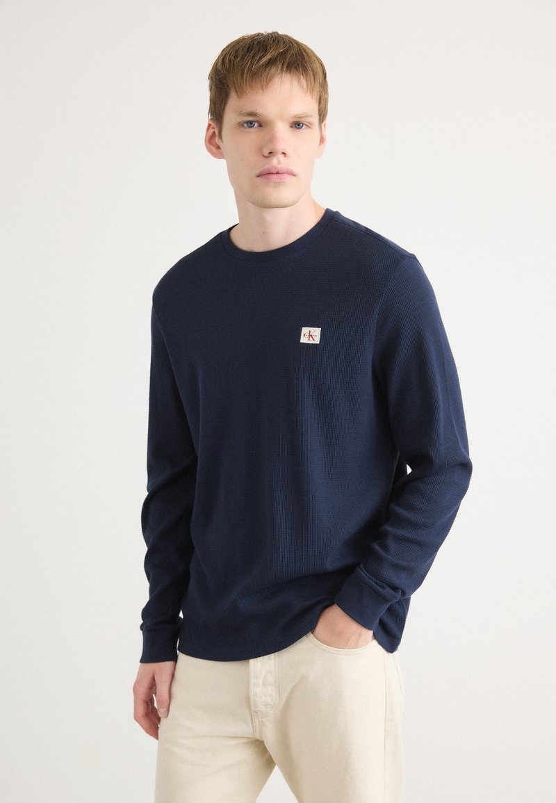 Navyblauwe lange mouwen shirt van een gestructureerde stof, ronde halslijn en een klein logopatch op de linker borst, gecombineerd met crèmekleurige broek.