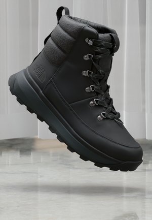 Botte noire imperméable avec un col rembourré, un devant à lacets et des accents texturés. Présente une semelle robuste et du matériel métallique.