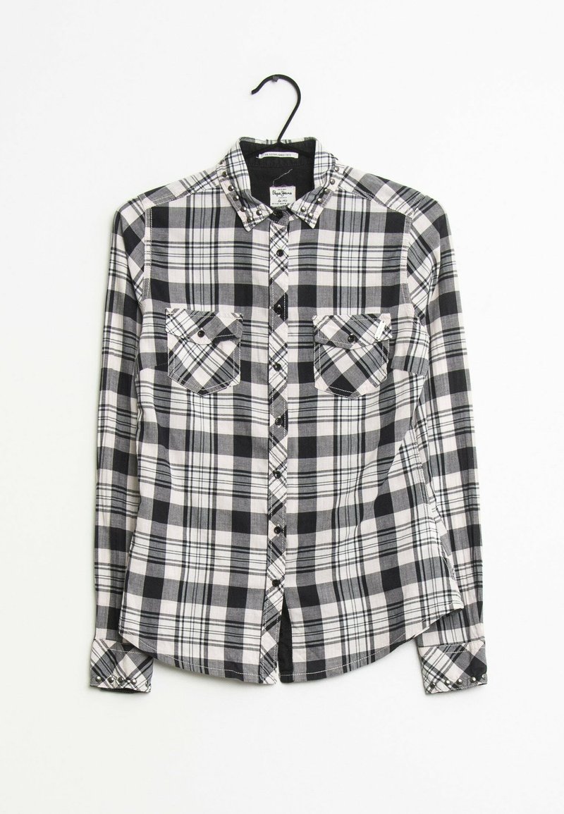 Chemise à carreaux noir et blanc à manches longues avec poches poitrine boutonnées suspendue sur un cintre noir sur fond blanc.