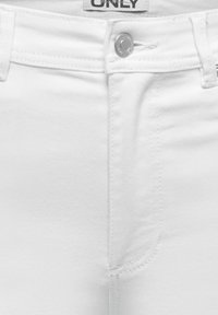 Jean en denim blanc en gros plan montrant la ceinture, le bouton argenté, les passants de ceinture, la braguette cousue et une étiquette de marque partielle indiquant "ONLY."