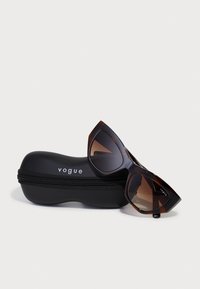 Ochelari de soare cu model din scoică de țestoasă, design unghiular de tip cat-eye și lentile degrade maro, așezați lângă o etui negru cu fermoar etichetat "vogue."