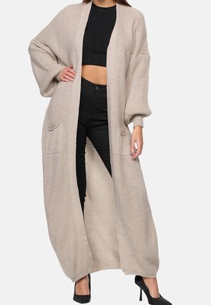 Femme portant un long cardigan beige en maille avec des poches, un crop top noir, un pantalon noir et des talons hauts noirs, debout avec la main sur la hanche.