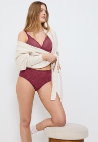 Burgundzki stanik z koronkowym wykończeniem i pasujące majtki z wysokim stanem, w połączeniu z jasnobeżowym swetrem, na neutralnym tle.