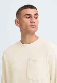 Camicia beige chiaro a maniche lunghe con scollo rotondo e tasca frontale. Il tessuto appare morbido e con una texture sottile.