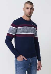 Suéter de punto azul marino con rayas horizontales rojas y blancas, con detalles texturizados en gris y cuello redondo. Logo de la marca en el hombro.