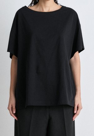 Kvinde iført en løs, sort T-shirt med korte ærmer og sorte bukser, stående mod en ensfarvet lys baggrund.