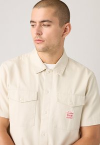 Short-sleeve beige shirt made of cotton with a button-down collar, two chest pockets, and a red label on the left pocket.