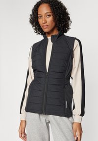 Čierna vypchávkovaná vesta s vysokým golierom a zipsom, s textúrovaným horizontálnym prešívaním a elastickými bočnými panelmi, kombinovaná s ľahkým vrchným oblečením.