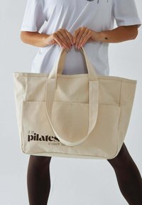 Beżowa torba tote z płótna, z dwoma mocnymi uchwytami, z przednią kieszenią i nadrukowanym tekstem "To jest czas na pilates" w kolorze brązowym. Gładka tekstura.