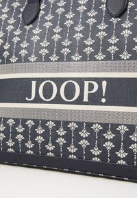 Torbica s mornarskim uzorkom i cvjetnim dizajnom, kontrastni bijeli logo "JOOP!" na teksturiranoj pozadini. Izrađena od izdržljivog materijala s strukturalnim oblikom.