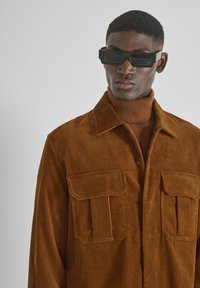 Bruine corduroy shirt jacket met twee voorkantzakken, knoopsluiting en een gemêleerde afwerking, gelaagd over een bijpassende coltrui.