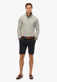 Lichtgrijze pullover met een kwart rits en marineblauwe accenten, gecombineerd met donkerblauwe shorts en bruine leren instappers; casual outfit.