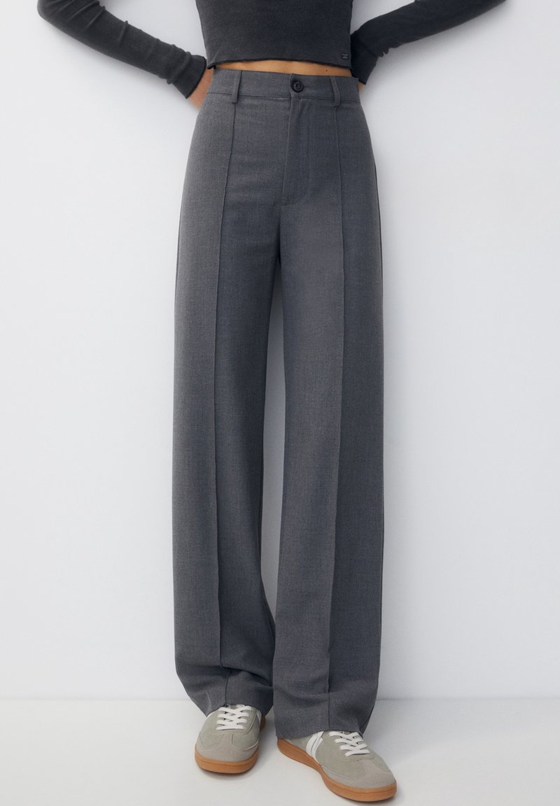 Personne portant un pantalon gris taille haute à jambes larges avec des plis à l'avant, associé à des baskets gris clair et un haut noir court à manches longues.