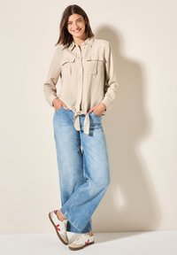 Camicia beige a maniche lunghe con due tasche frontali, annodata in vita, abbinata a jeans a gamba larga azzurri e sneaker beige con dettagli rossi.