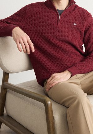 Mężczyzna siedzący na beżowym, tapicerowanym, drewnianym krześle, ubrany w teksturowany, ciemnoczerwony sweter na zamek i beżowe spodnie, z rękami spoczywającymi na kolanach i podłokietniku.