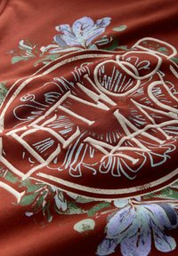 Tissu couleur rouille avec le mot « Renewed » brodé en blanc, entouré d'un motif floral en bleu clair et vert.