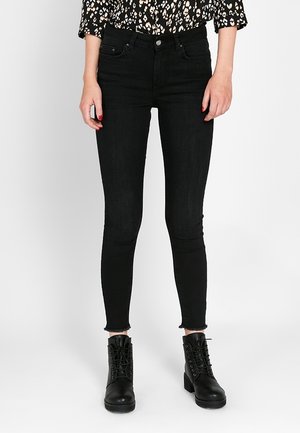 Femme portant un jean skinny noir avec des ourlets effilochés, des bottines noires à lacets et un haut noir à manches longues à motifs, debout sur un fond blanc.