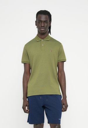 Vyras stovi, vilki alyvuogių žalumo polo marškinėlį ir tamsiai mėlynas šortus prieš paprastą baltą foną.