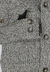 Grau-weißer Strickpullover mit einem strukturierten Muster, fünf dunklen Knöpfen und einer Seitentasche. Das Material wirkt weich und warm.