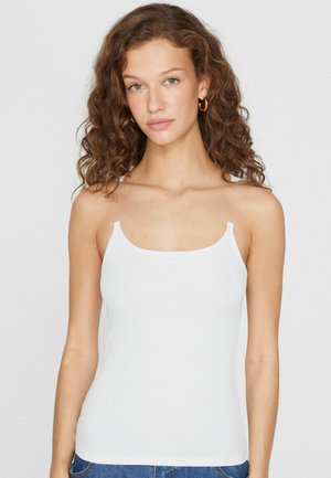 Joven con cabello rizado, lleva pendientes de aro dorados y una blusa blanca sin mangas, posando frente a un fondo claro y sencillo.