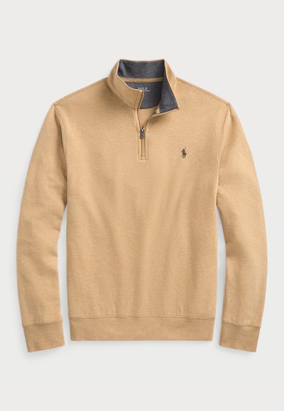 Beige pullover med en fjärdedelsdragkedja, med en mörkblå inre fodring och en liten broderad logotyp på bröstet. Mjuk textur.