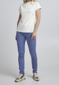 Bílý top s krátkým rukávem a plisováním na krku, doplněný světlými modrými slim-fit kalhotami. Modelka má na sobě béžové a šedé tenisky s texturovanými detaily.