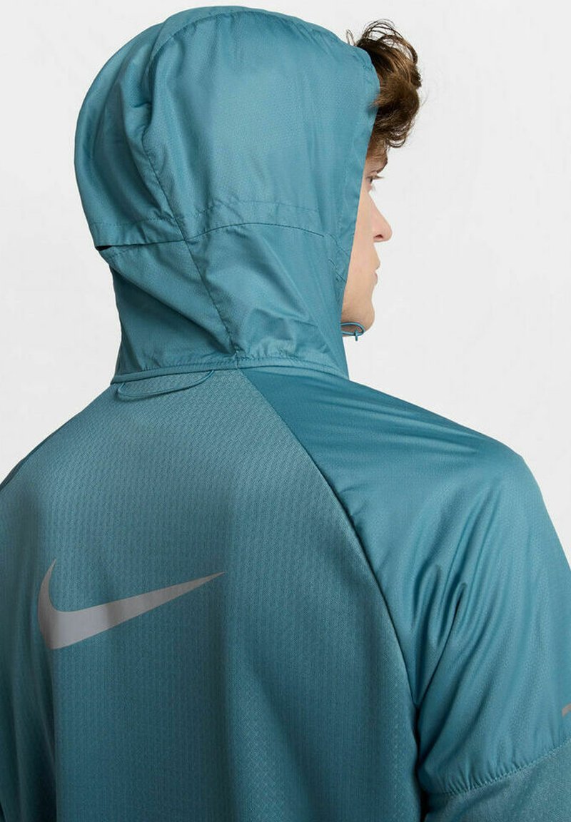 Veste à capuche teal en tissu texturé, avec un logo Nike réfléchissant au dos. Design léger avec une forme profilée.