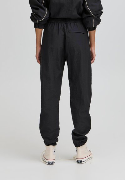 Schwarze, leichte Jogginghosen mit einem elastischen Bund und Bündchen. Verfügt über eine Gesäßtasche und einen strukturierten Stoff. Getragen mit weißen Sneakers.