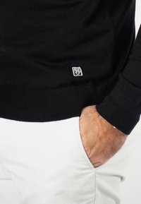 Pull en tricot noir avec un ourlet côtelé, présentant un petit patch logo en bas à gauche. Porté avec un pantalon blanc, mettant en valeur un tissu texturé.
