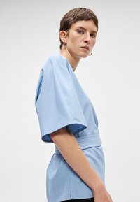 Ανοιχτό μπλε μπλούζα με φαρδιά κοντά μανίκια, ελαστική μέση και ομαλή υφή. Ο σχεδιασμός διαθέτει μια διακριτική γυαλάδα και ελάχιστες λεπτομέρειες.