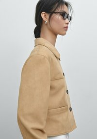 Tan suede jas met een kraag, knoopsluiting en zijzakken. Model draagt zwarte zonnebril. Zachte textuur en zorgvuldig gesneden pasvorm.