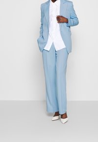 Completo su misura azzurro chiaro composto da una giacca monopetto e pantaloni a gamba larga, abbinato a una camicia bianca con bottoni e tacchi a punta bianchi.