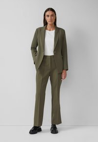 Blazer ajustado verde oliva y pantalones a juego, combinados con una camisa blanca. Presenta un solo botón y un corte elegante, usado con zapatos negros.