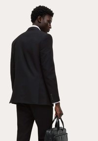 Czarny garnitur o dopasowanym kroju, biała koszula widoczna u mankietów, trzymana w czarnej skórzanej teczce. Gładka faktura, minimalistyczny design.