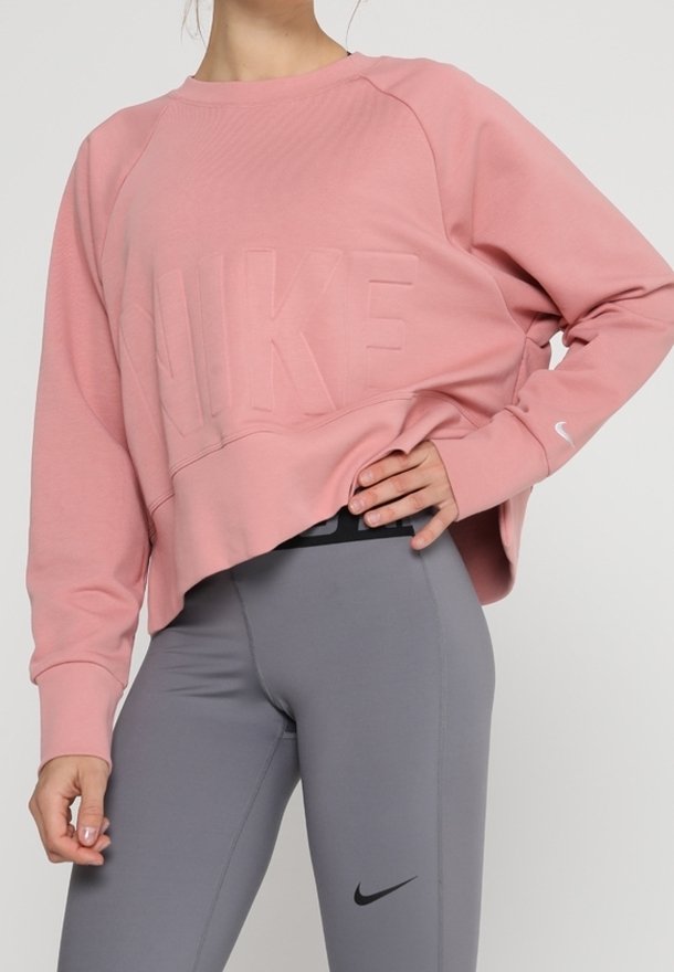 Personne portant un sweat-shirt Nike rose et un legging gris Nike taille haute, debout avec une main sur la hanche.