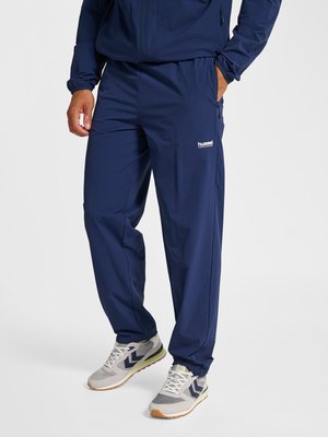 Man met donkerblauwe trainingsbroek en jas met wit logo, staand met één hand in de zak en grijze sneakers met witte veters.