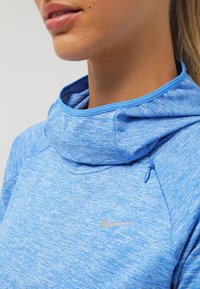 Hellblauer Kapuzenpullover mit strukturierter Optik, ausgestattet mit hohem Kragen, kleinem Reißverschlusszug und einem silbernen Nike-Logo auf der Brust.
