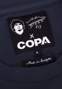 Zwarte label met een witte grafische afbeelding van een persoon met krullend haar, "X COPA", maat "S", en tekst "Made in Europe" op een marineblauwe stof achtergrond.