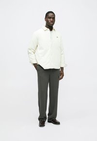 Chaqueta beige claro abotonada con textura suave, usada sobre una camisa blanca, combinada con pantalones grises y zapatos de cuero oscuro, posando sobre un fondo neutro.