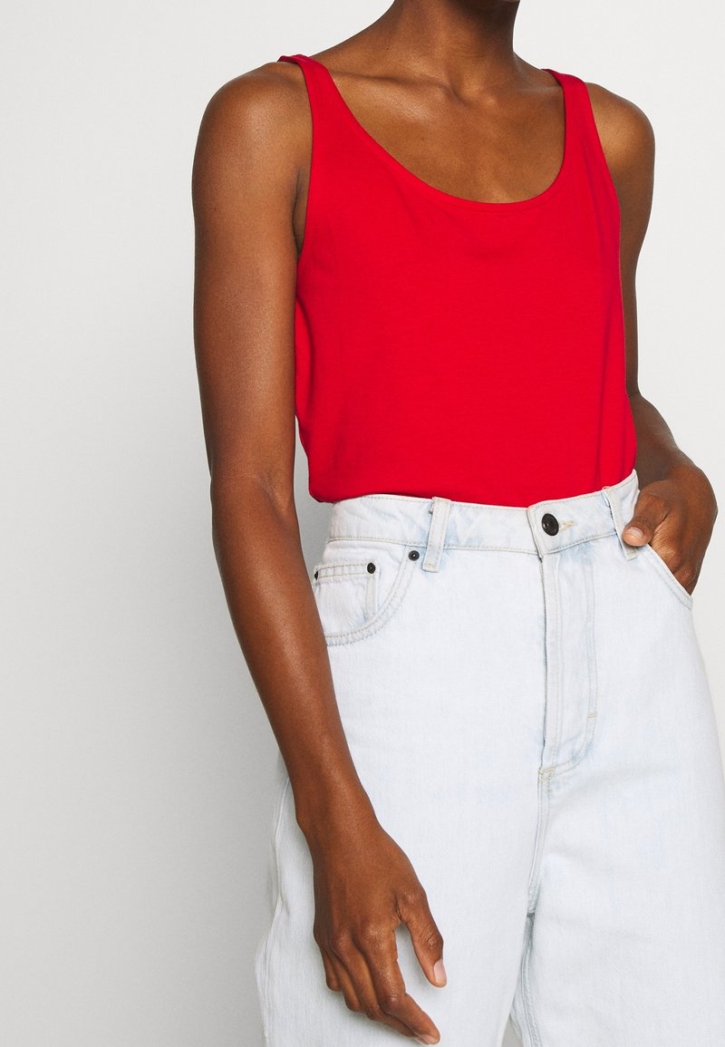 Haut rouge sans manches associé à un jean en denim bleu clair taille haute, une main dans la poche et l'autre le long du corps.