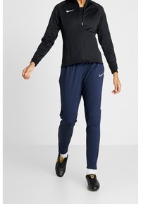 Svart Nike zip-up jacka tillsammans med marinblå träningsbyxor. Jackan har långa ärmar och en figurnära design; byxorna har en avsmalnande form med vita detaljer.
