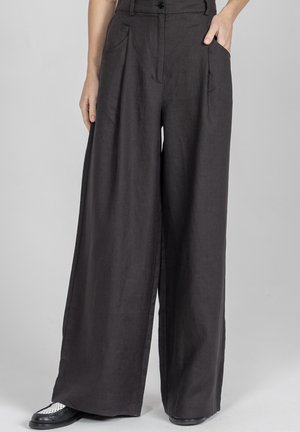 Ciemnoszare spodnie o wysokim stanie i szerokich nogawkach z plisami z przodu, bocznymi kieszeniami oraz czarno-białe buty na osobie stojącej na jednolitym tle.